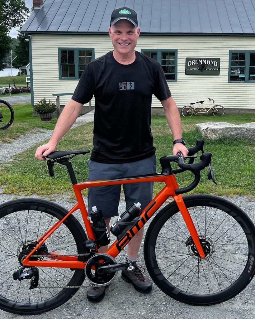 Darryl Jelley standing next to an orange BMC road bike outside Drummond custom bike shop Enfield, NH.